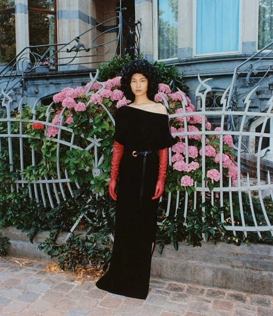Asian woman in a long black dress with red gloves poses in front of a building with wrought iron and pink hydrangea bushes.