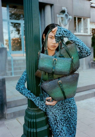 Fashion shot of a woman with long dark hair, wearing a patterned dress, holding 3 green and black woven handbags near a green lamppost.