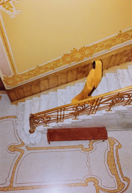 High angle view of person in yellow gown ascending marble stairs with ornate gold railing and floor detail in an opulent setting.