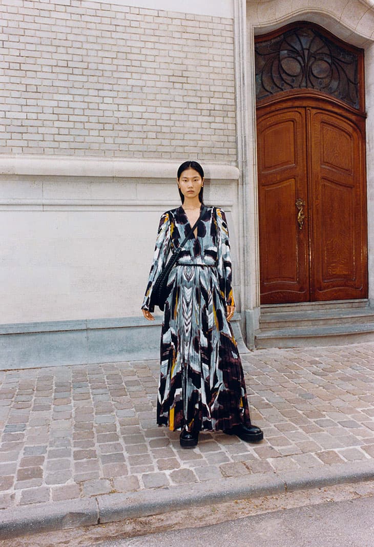 An Asian woman stands outdoors in an abstract-print, long-sleeved maxi dress, black shoulder bag, and chunky-heeled boots, on cobblestone.