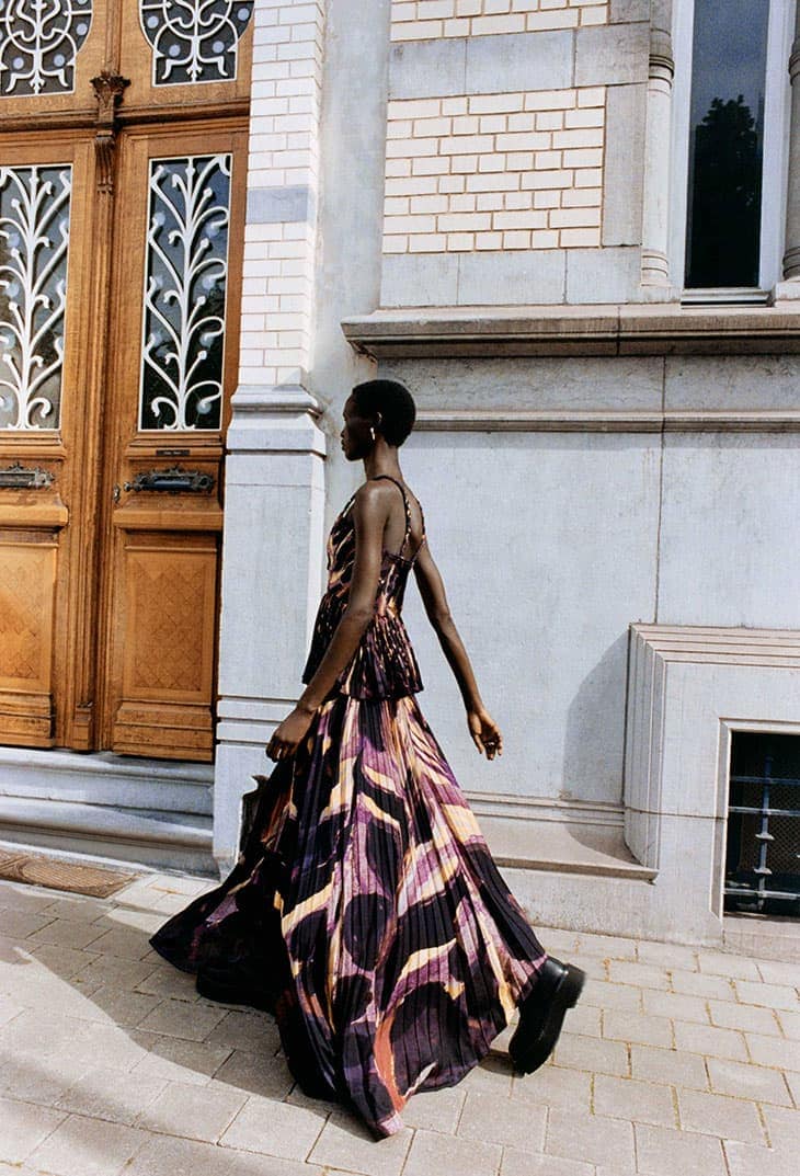 Fashion shot of an African-American woman in a patterned dress walking on a sidewalk next to a building with a wooden door.