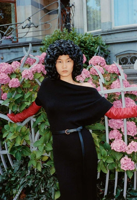 Asian woman in black dress and red gloves poses near hydrangea bush and wrought-iron gate in an outdoor fashion setting.