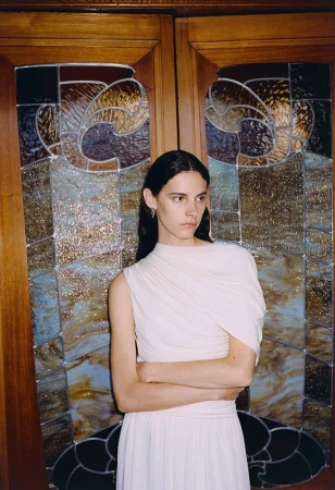 Portrait of a woman in a cream dress standing in front of a stained glass door with her arms crossed in a studio setting.