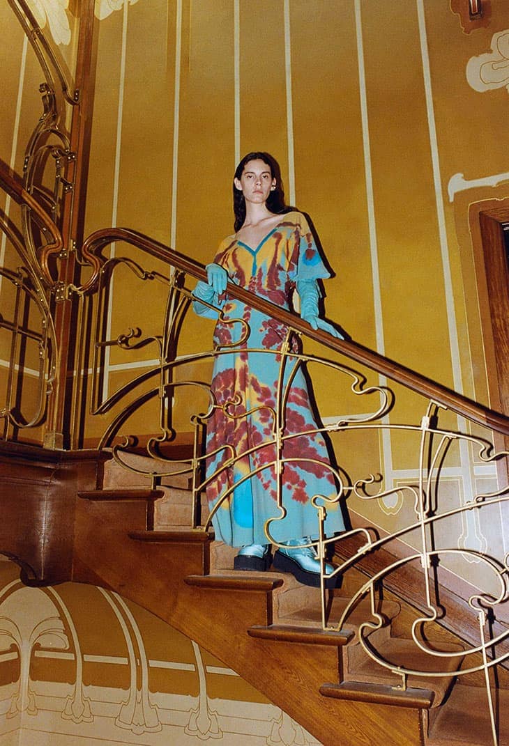 A woman poses in a tie-dye dress on wooden stairs with decorative gold railings against a yellow wall with matching gold details.