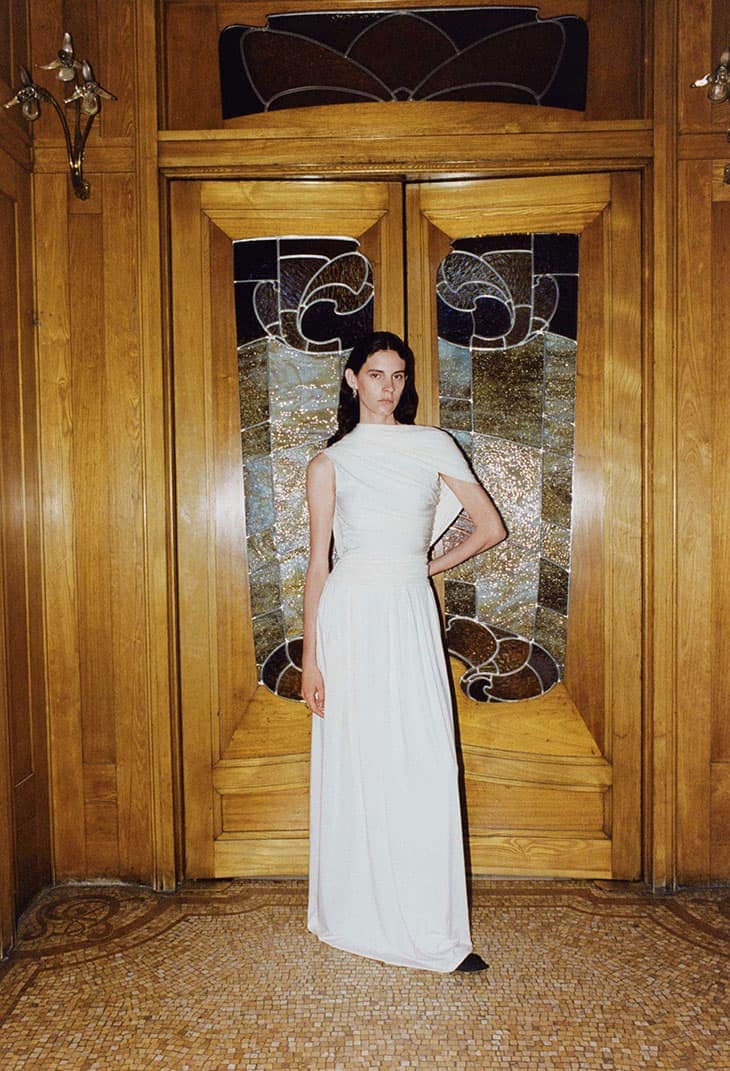 A woman in a white dress stands in front of a wooden door with glass inserts. The photo has an editorial fashion aesthetic.
