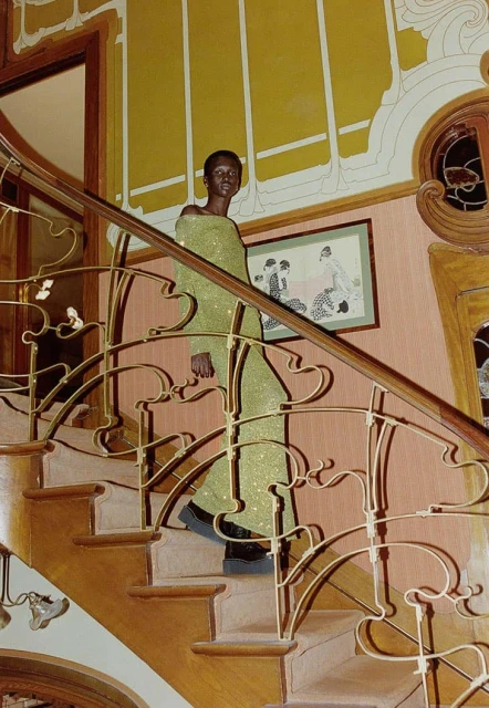 Adult woman with dark skin and a shaved head poses on an indoor staircase wearing an off-shoulder, floor-length chartreuse dress.