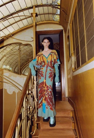 A model poses on an ornate staircase wearing a patterned dress and gloves in a fashion editorial style shot with architectural details.
