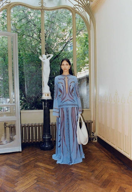 Indoor fashion shot of a woman in a blue patterned dress holding a white purse, standing in front of a window.