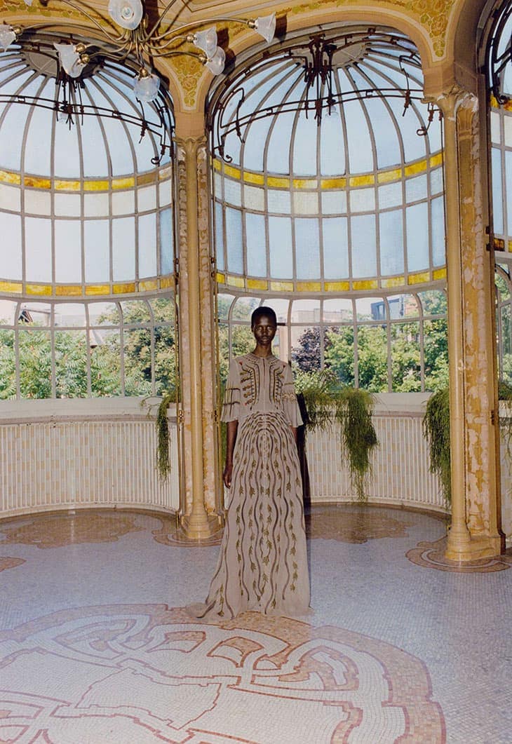 Woman poses in a beige dress with decorative embroidery in an ornate, light-filled hall featuring decorative columns and a mosaic floor.