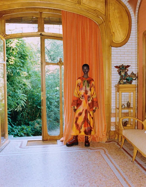 Indoor fashion shot of a Black female model in a colorful dress, standing in a room with a large open window and peach curtains.