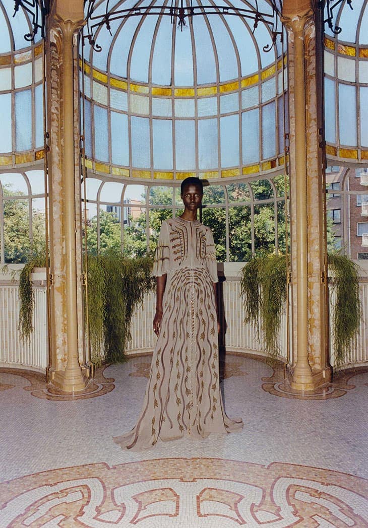 Woman in a beige gown with abstract design posing in a sunroom with a tiled floor and large arched windows with greenery and outdoor view.