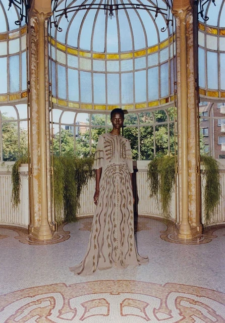 Woman in a beige gown with abstract design posing in a sunroom with a tiled floor and large arched windows with greenery and outdoor view.