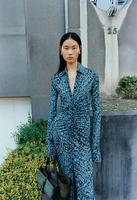 Elegant Asian woman in long-sleeve blue-patterned dress holds bag outdoors by gray building with green plants, fashion editorial.