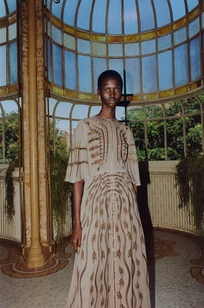Woman in long dress with brown geometric pattern poses in front of an architectural greenhouse structure filled with greenery.