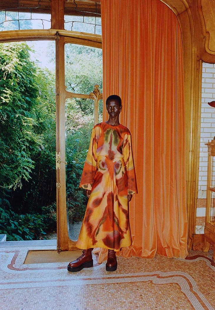 An African woman stands in a colorful dress inside an ornate building next to a large window that frames lush greenery outdoors.