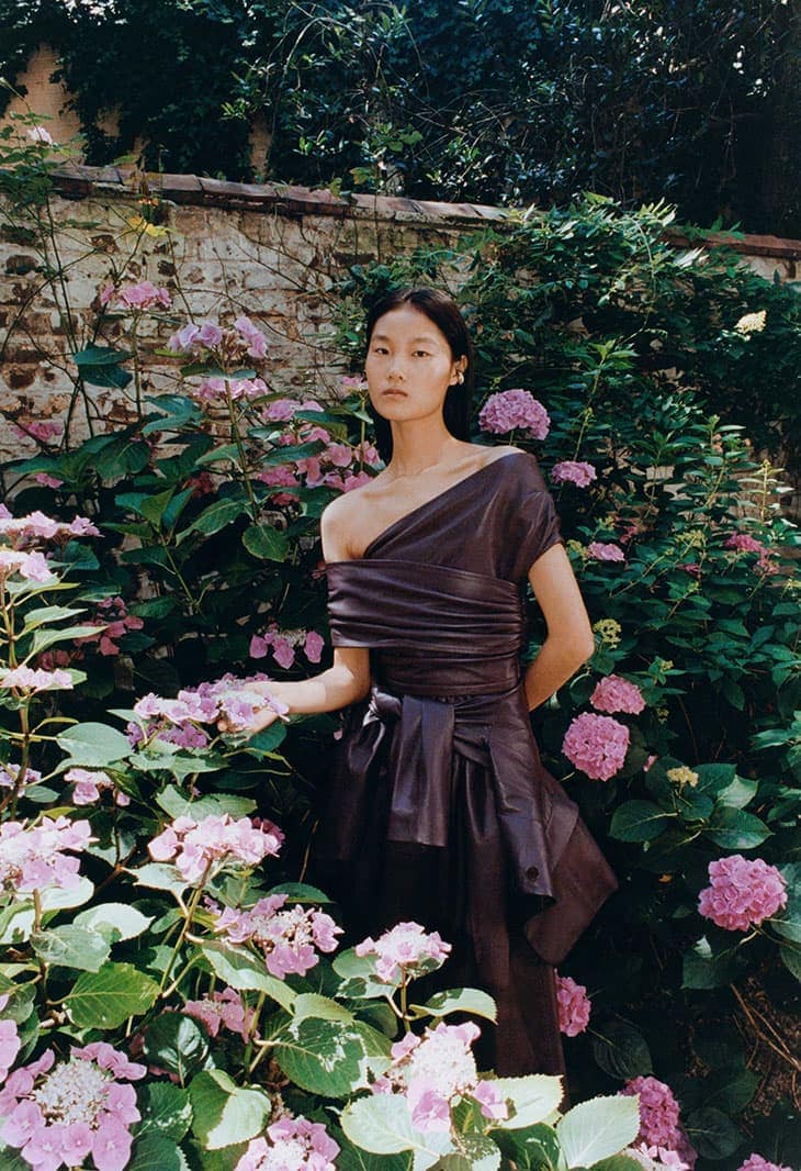 Asian woman in an elegant brown one-shoulder dress posing amongst blooming pink hydrangea flowers in a lush garden setting.