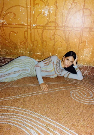 Woman in a patterned dress lies on a mosaic floor against an ornate, distressed wall in an indoor setting with natural light.