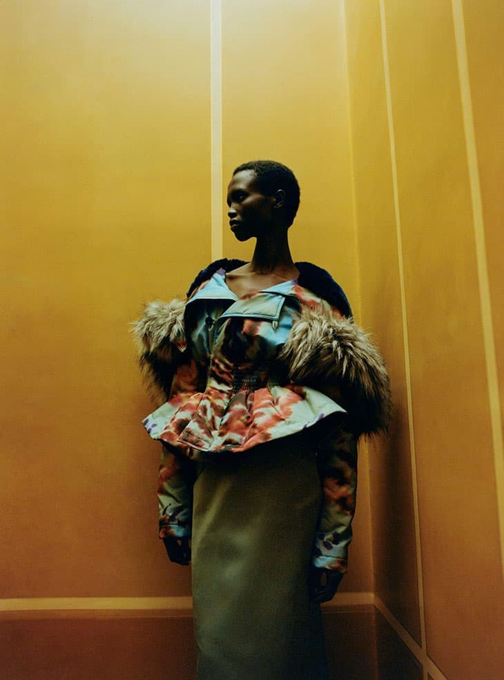 Fashion photo of a Black woman in a patterned fur-trimmed jacket and a long skirt, posing against a yellowish-orange wall with vertical lines.