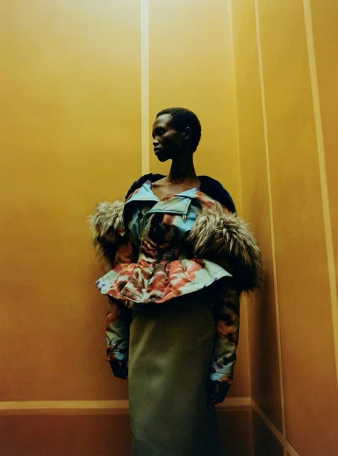 Fashion photo of a Black woman in a patterned fur-trimmed jacket and a long skirt, posing against a yellowish-orange wall with vertical lines.