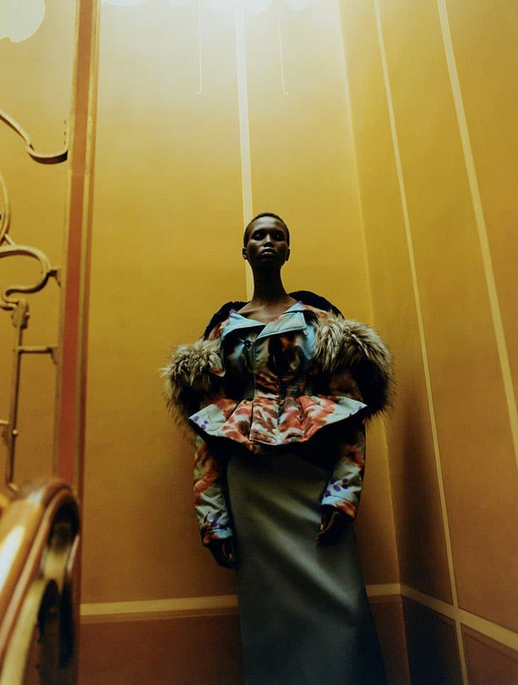 Fashion shot of a dark-skinned woman in a unique patterned and fur-trimmed jacket, standing in a warm-toned interior with vertical lines.