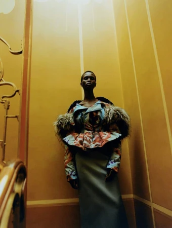 Fashion shot of a dark-skinned woman in a unique patterned and fur-trimmed jacket, standing in a warm-toned interior with vertical lines.