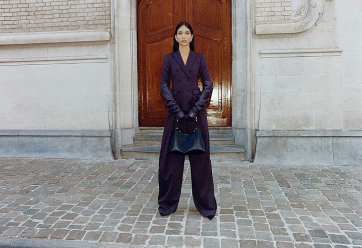 Elegant woman in purple suit and long leather gloves stands outdoors holding a black bag, posed in front of a weathered building and door.