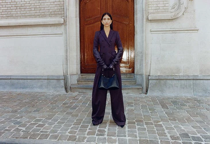 Elegant woman in purple suit and long leather gloves stands outdoors holding a black bag, posed in front of a weathered building and door.
