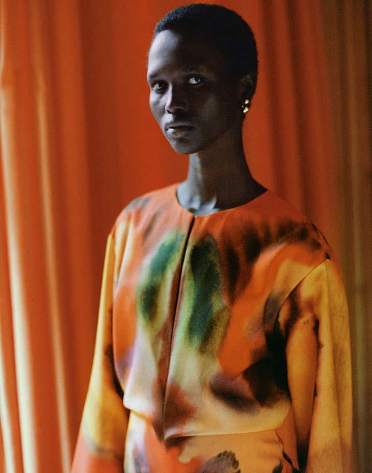 Close-up portrait of a Black female model wearing a colorful orange, yellow, and green patterned top in front of an orange curtain.