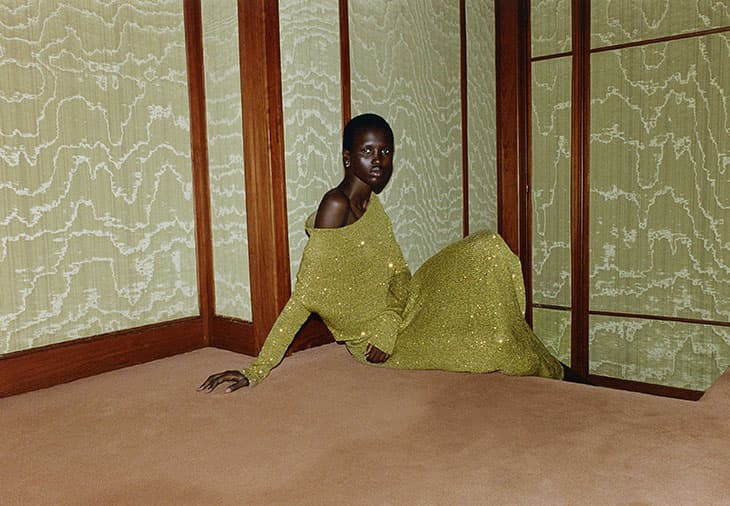 Elegant black woman in gold, glittering long-sleeve dress, sitting gracefully indoors against patterned wallpaper and dark-wood frame.
