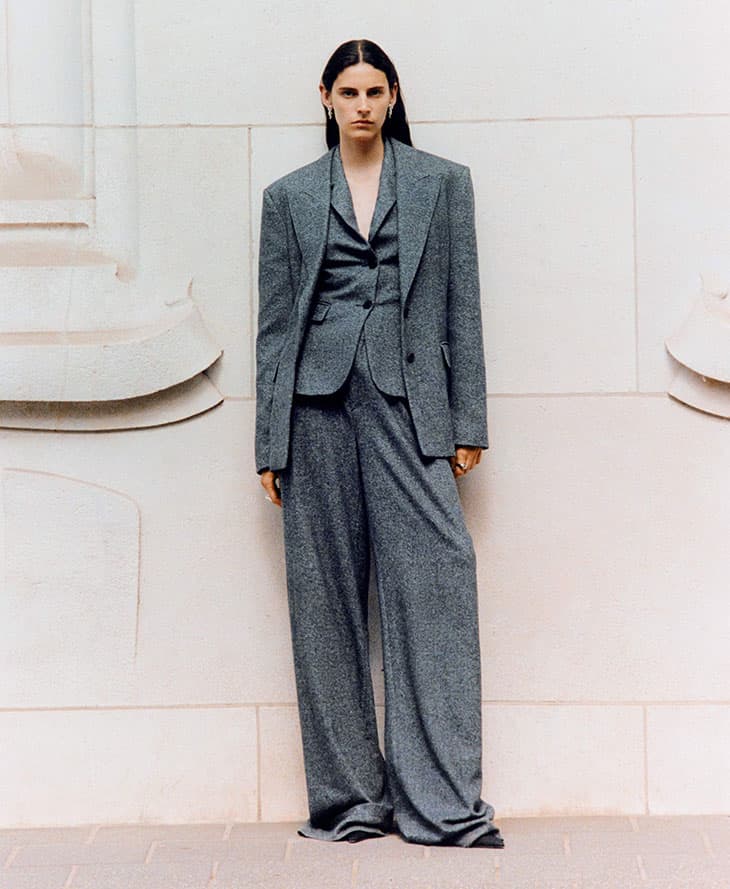 Woman poses in an oversized grey suit, including jacket, vest, and wide-leg pants, in front of a light-colored architectural wall.