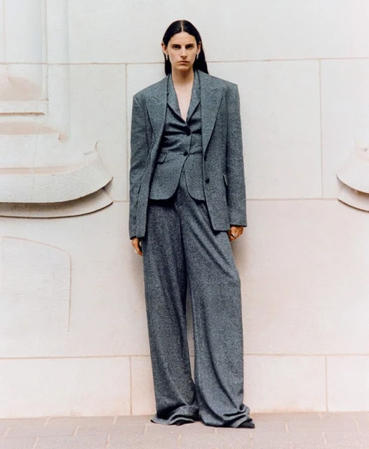 Woman poses in an oversized grey suit, including jacket, vest, and wide-leg pants, in front of a light-colored architectural wall.