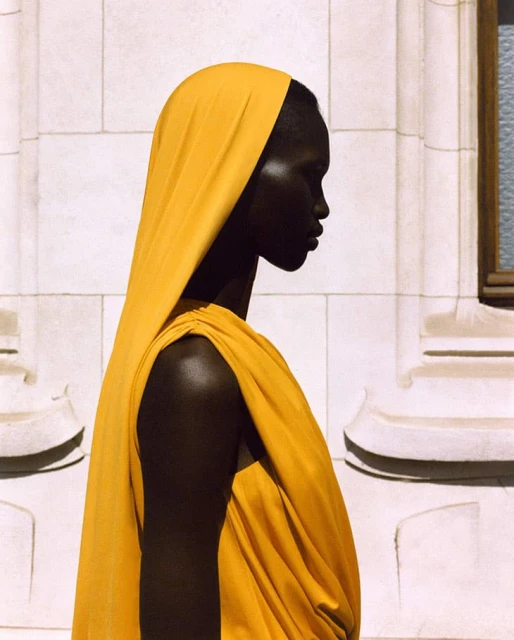 Profile portrait of an African-descent adult woman wearing a yellow headscarf and draped garment against a light-colored building facade.