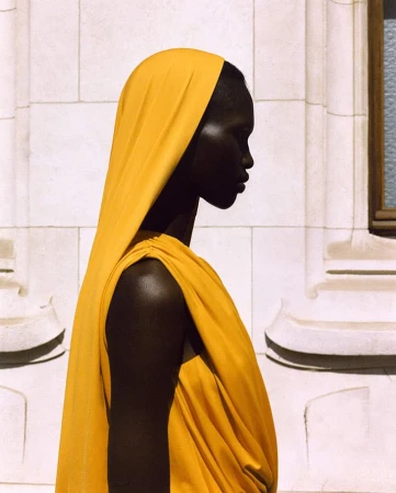 Profile portrait of an African-descent adult woman wearing a yellow headscarf and draped garment against a light-colored building facade.