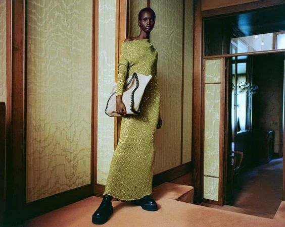 A Black female model in a long, shimmering green dress holds a white tote bag indoors, standing on a pink step in front of a doorway.