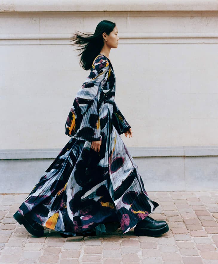 A slender female fashion model with long black hair walks outside. She wears a long, pleated dress with a unique artistic print.