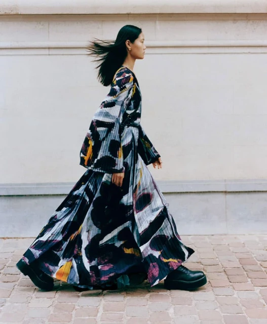 A slender female fashion model with long black hair walks outside. She wears a long, pleated dress with a unique artistic print.