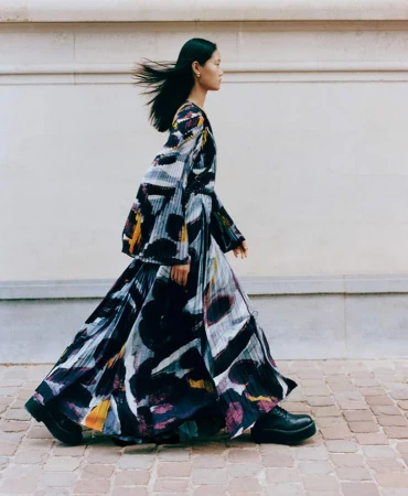 A slender female fashion model with long black hair walks outside. She wears a long, pleated dress with a unique artistic print.