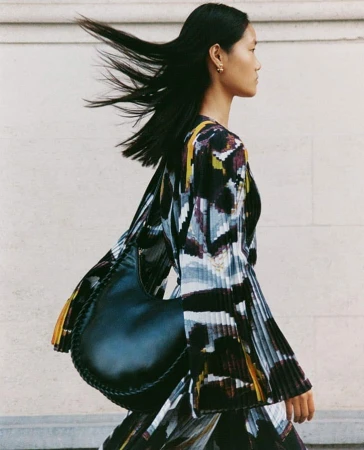 Fashionable Asian woman with a black handbag, striking black hair, and an abstract print dress stands against a neutral-toned wall.