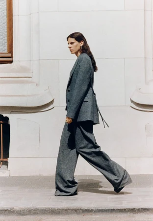 A woman with long dark hair wears a grey wool trouser suit while walking past a light stone building wall.