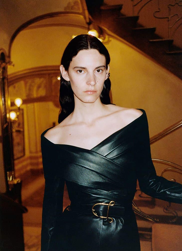 Woman in a dark, off-the-shoulder leather dress with a wide belt and gold buckle, posing indoors against a warm-toned stairwell background.