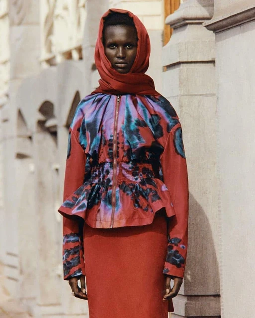 Adult female in a red headscarf and skirt suit poses against a textured, off-white stone facade. Medium shot, outdoor setting.