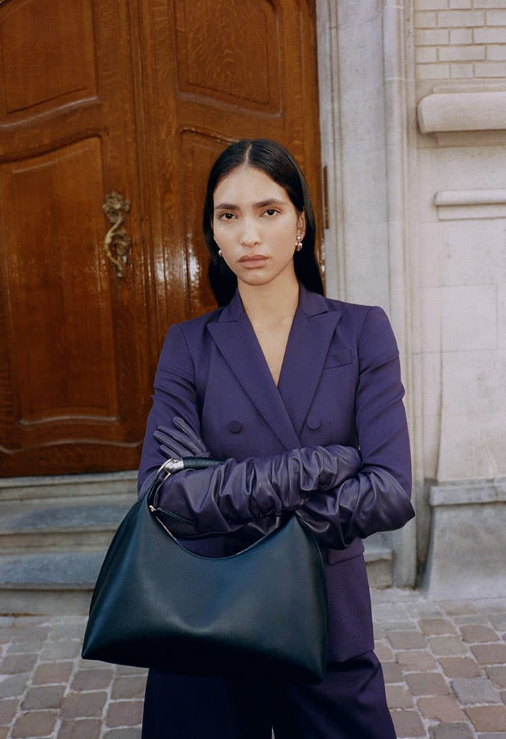 Woman in purple suit and gloves holds dark-green structured handbag against wood door and stone pavement in daylight outdoor setting.