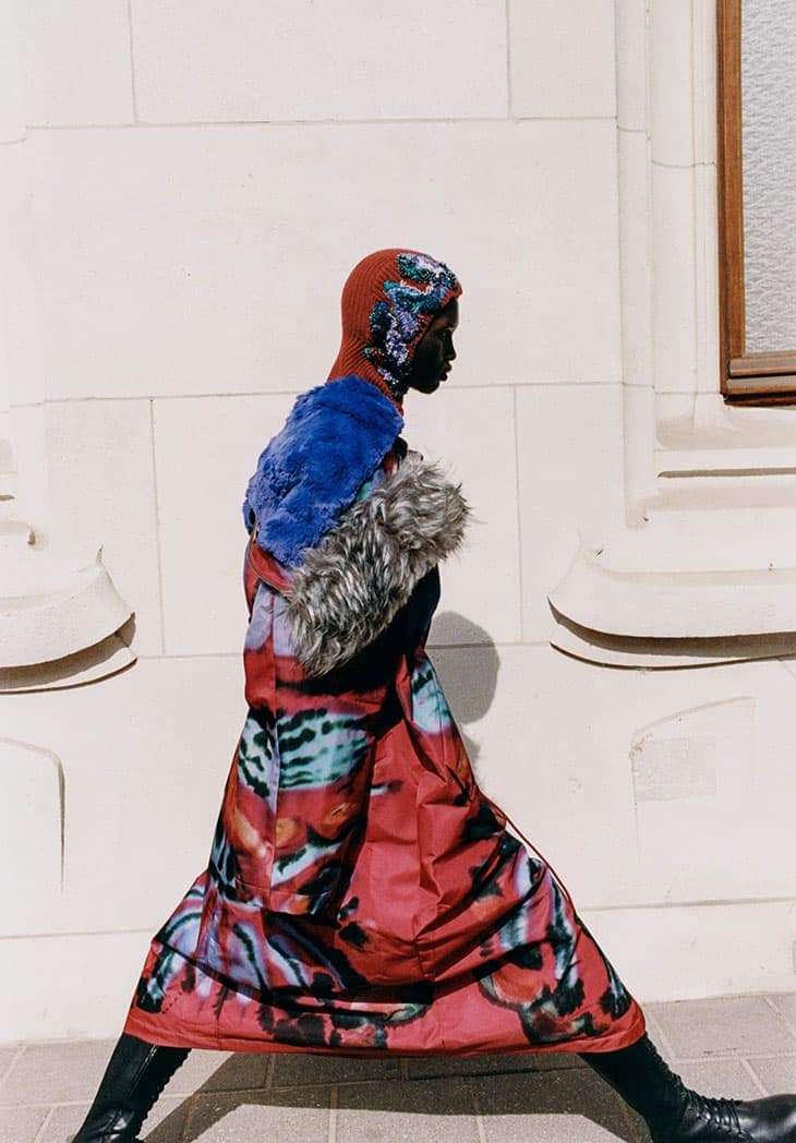 A model walks in an editorial fashion shot, wearing a multi-colored coat, cap and boots against a light wall with a framed window.