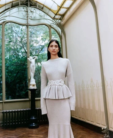 Indoor shot of a woman in a matching sweater and skirt, posing near a sculpture in a naturally lit interior space.