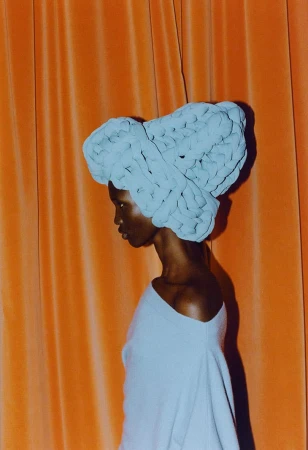 Profile view of Black woman wearing a light-blue, textured head wrap, against a draped orange curtain backdrop in a studio setting.