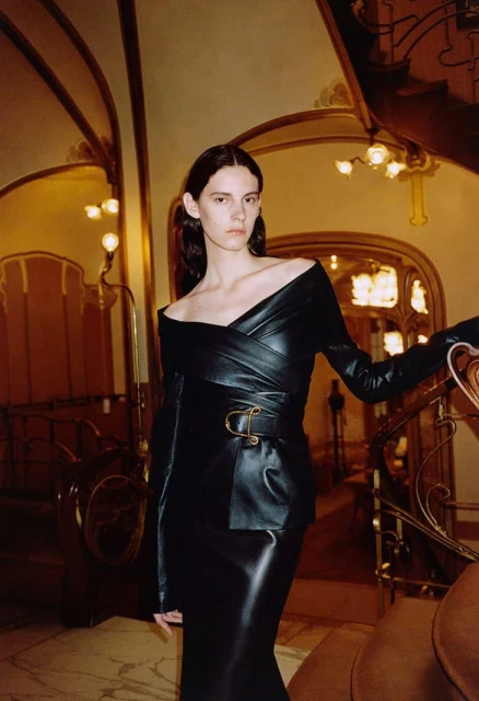 Elegant adult female poses on stairs in a black off-the-shoulder leather dress with a gold buckle. Architectural background.