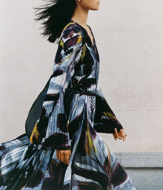 Fashion shot of a woman with dark hair wearing a long-sleeve dress with a flowing skirt and abstract print, walking against a light-colored wall.