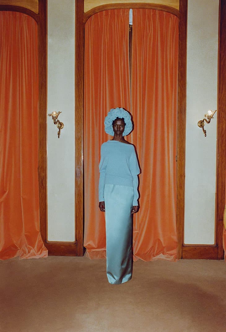 Elegant dark-skinned woman in light blue sweater, floor-length skirt, and floral hat posing indoors against peach curtains and beige walls.