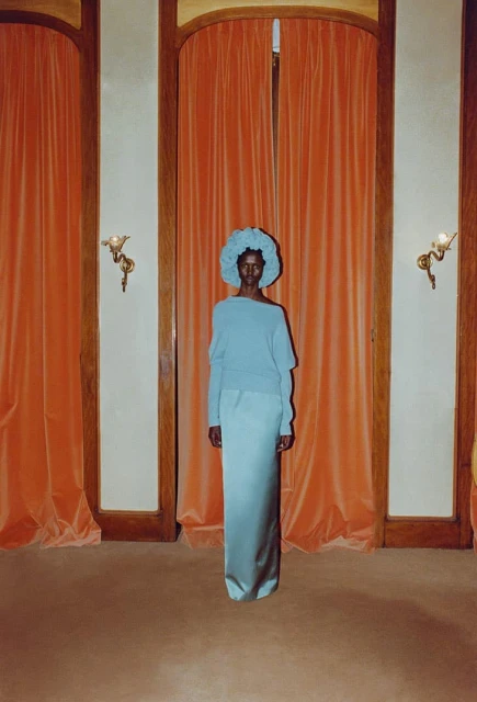 Elegant dark-skinned woman in light blue sweater, floor-length skirt, and floral hat posing indoors against peach curtains and beige walls.