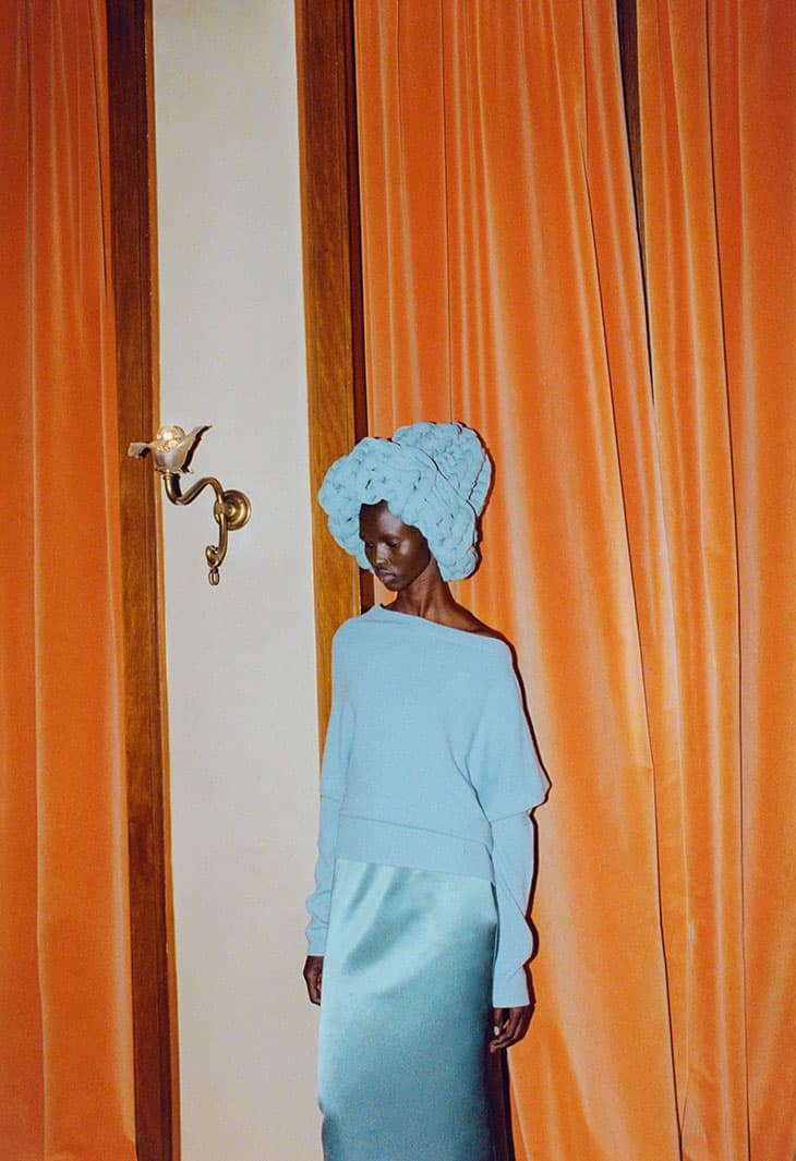 Fashion shot of a Black woman in a blue sweater and skirt with a matching blue head covering, posing indoors against a warm orange curtain.